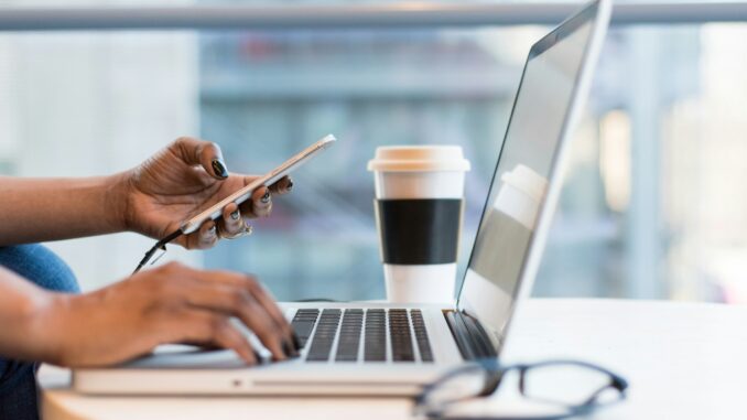 Photo of a person using their mobile smartphone while at a computer to illustrate the launch of OpenAI Operator that promises to democratise agentic AI by bringing agents into the browser.